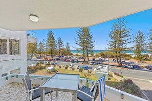 Een balkon met een glazen tafel en stoelen, met uitzicht op een parkeerplaats en een strand met bomen.