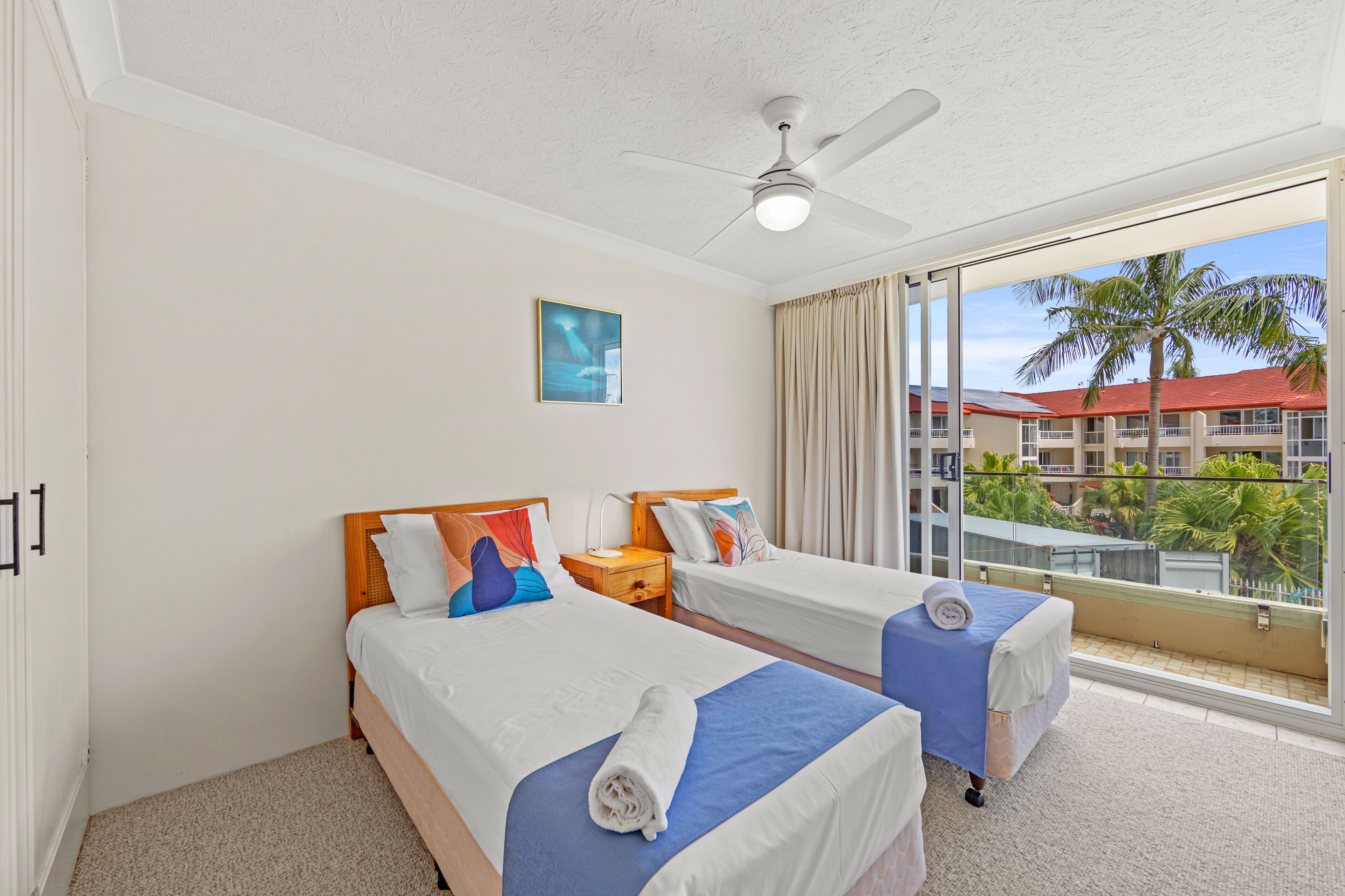Habitación de hotel con dos camas, un ventilador de techo, un ventanal con vistas a edificios y palmeras, y un cuadro en la pared.