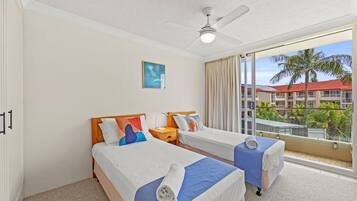 A hotel room with two beds, a ceiling fan, a large window with a view of buildings and palm trees, and a painting on the wall.