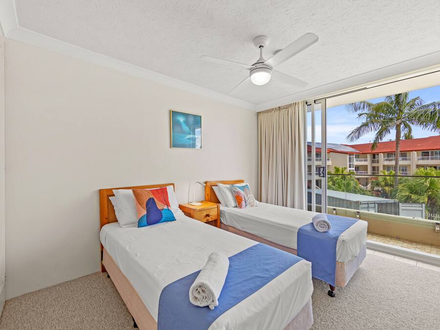 Habitación de hotel con dos camas, un ventilador de techo, un ventanal con vistas a edificios y palmeras, y un cuadro en la pared.