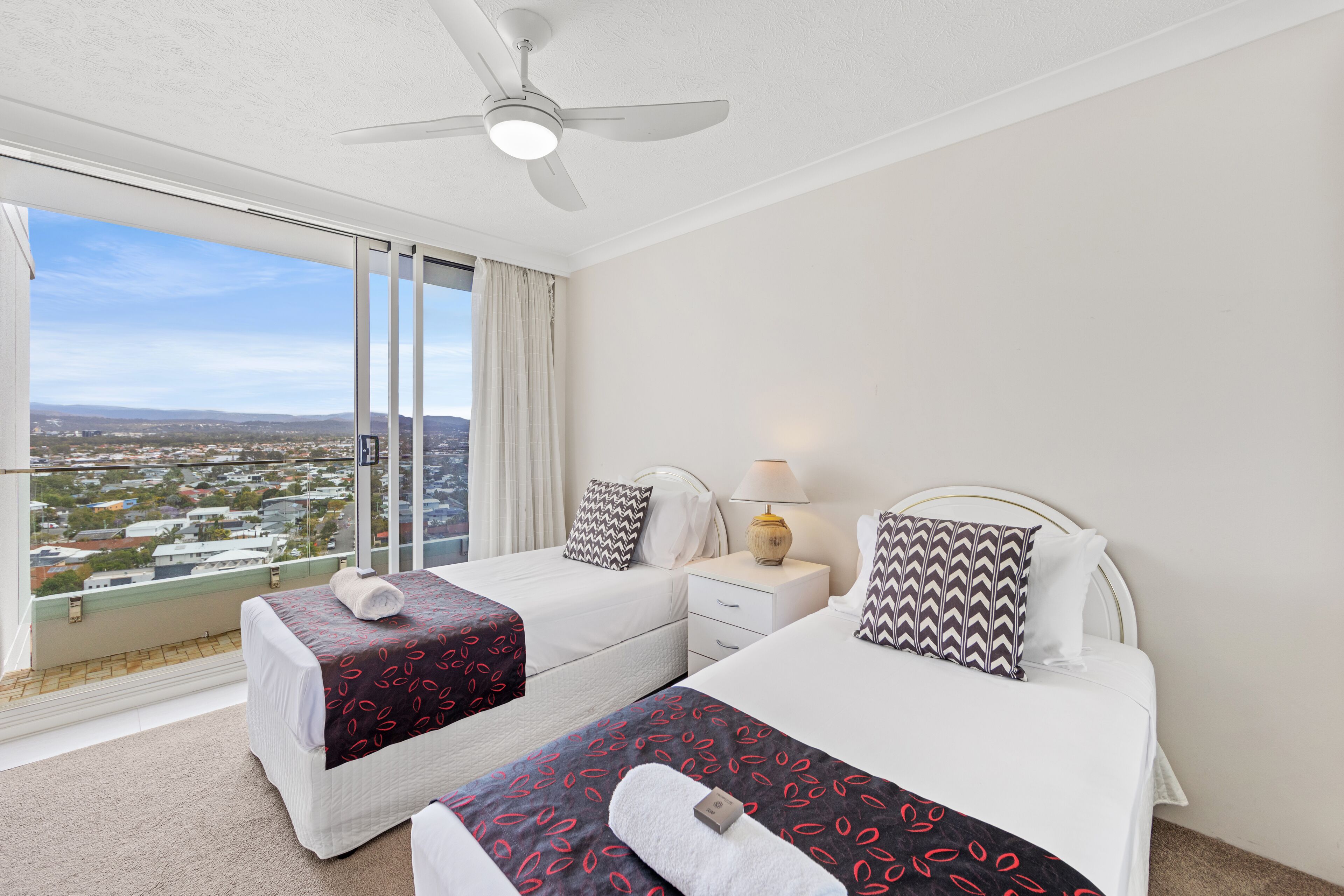 A hotel room with two beds, a ceiling fan, a nightstand with a lamp, and a large window offering a city view.