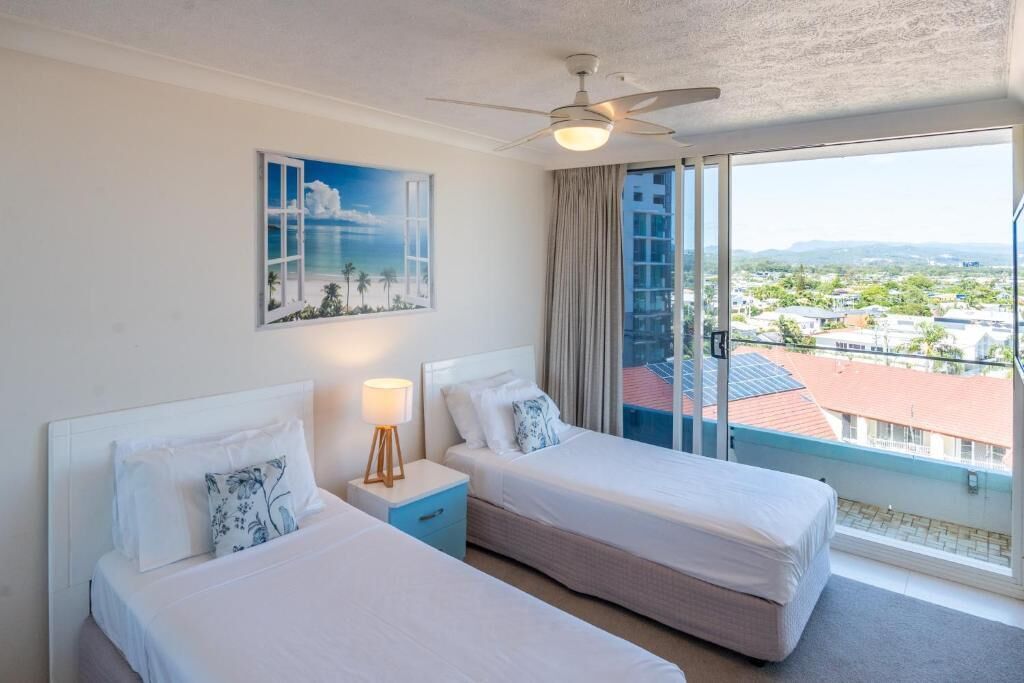 Habitación de hotel con dos camas, ventilador de techo, ventana grande con vista a la ciudad y un cuadro en la pared.