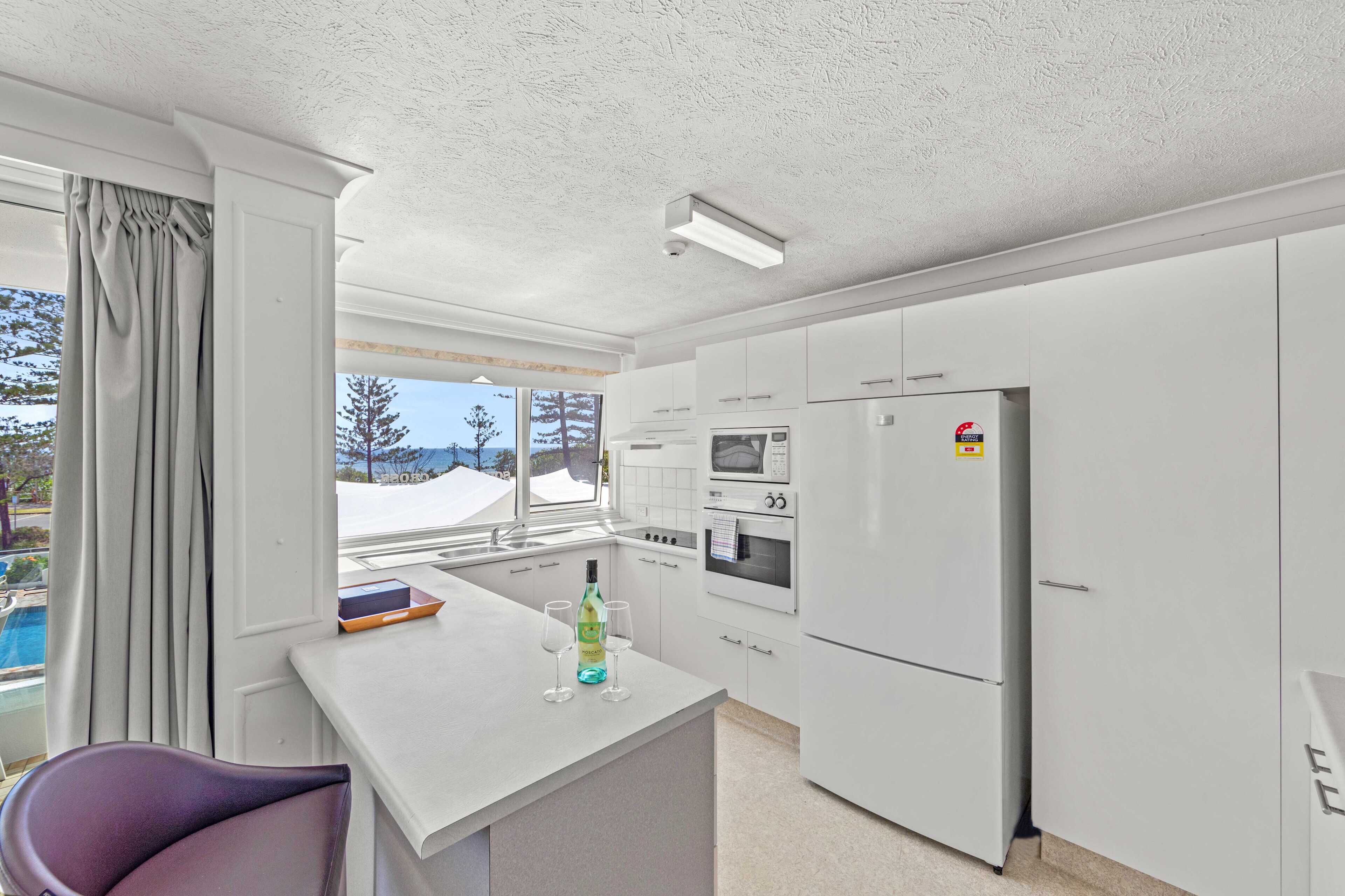 Una cocina moderna con armarios blancos, un refrigerador, un horno y un microondas. Hay una ventana con vistas a árboles y una piscina.