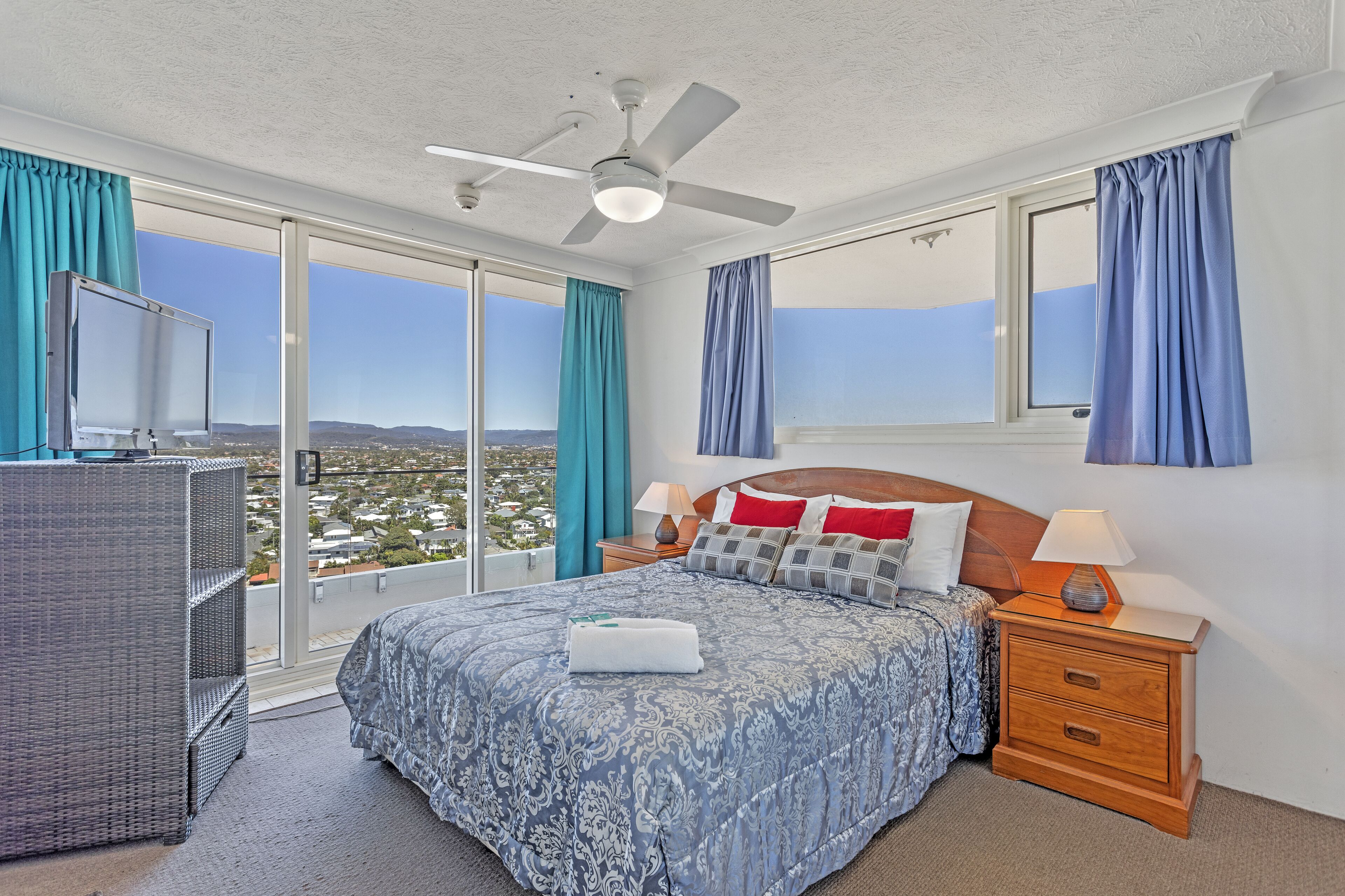 A bedroom with a large bed, a nightstand, a dresser, and a view of the city through a large window.