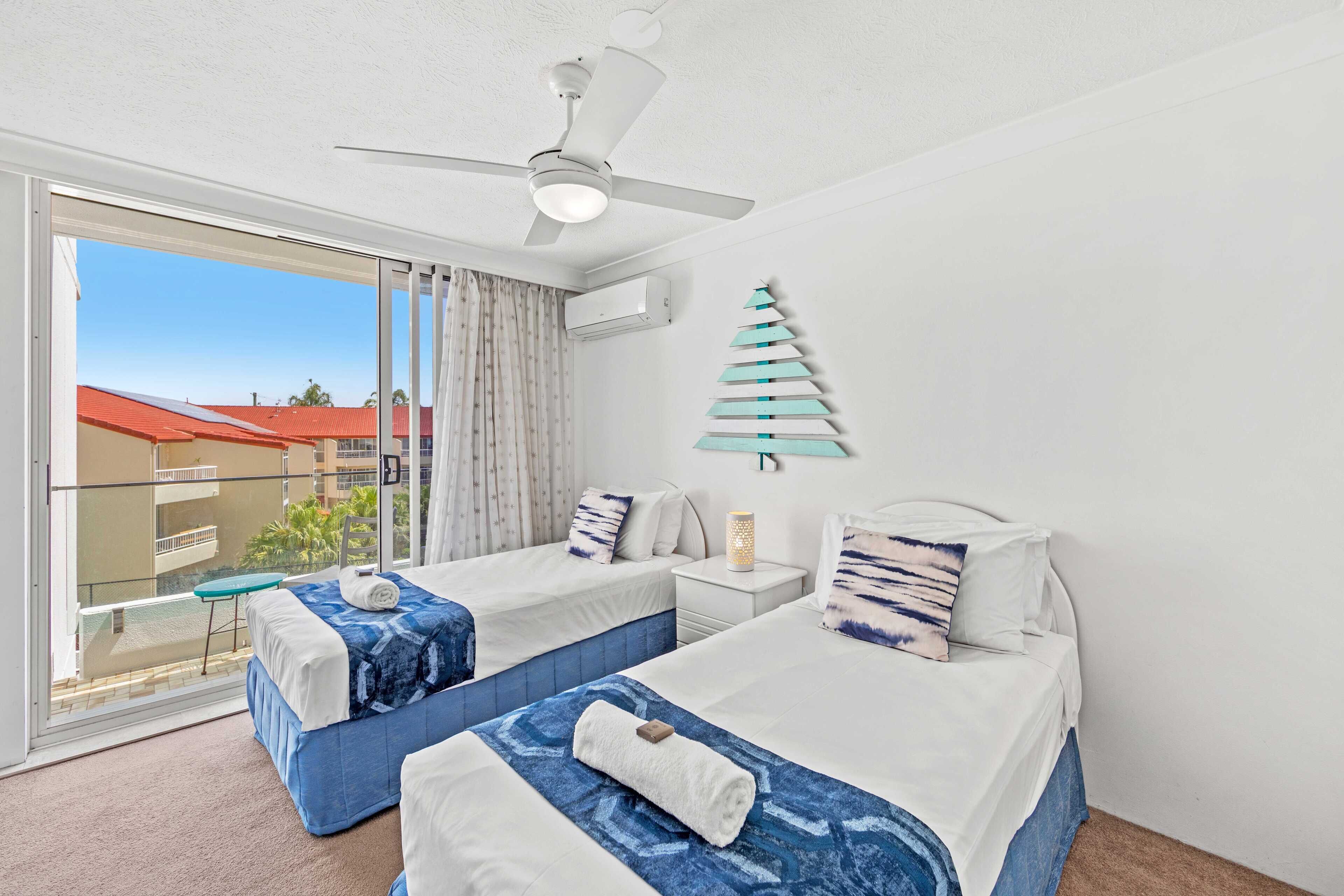 A hotel room with two beds, a ceiling fan, a decorative wall piece, and a balcony with a view of buildings.