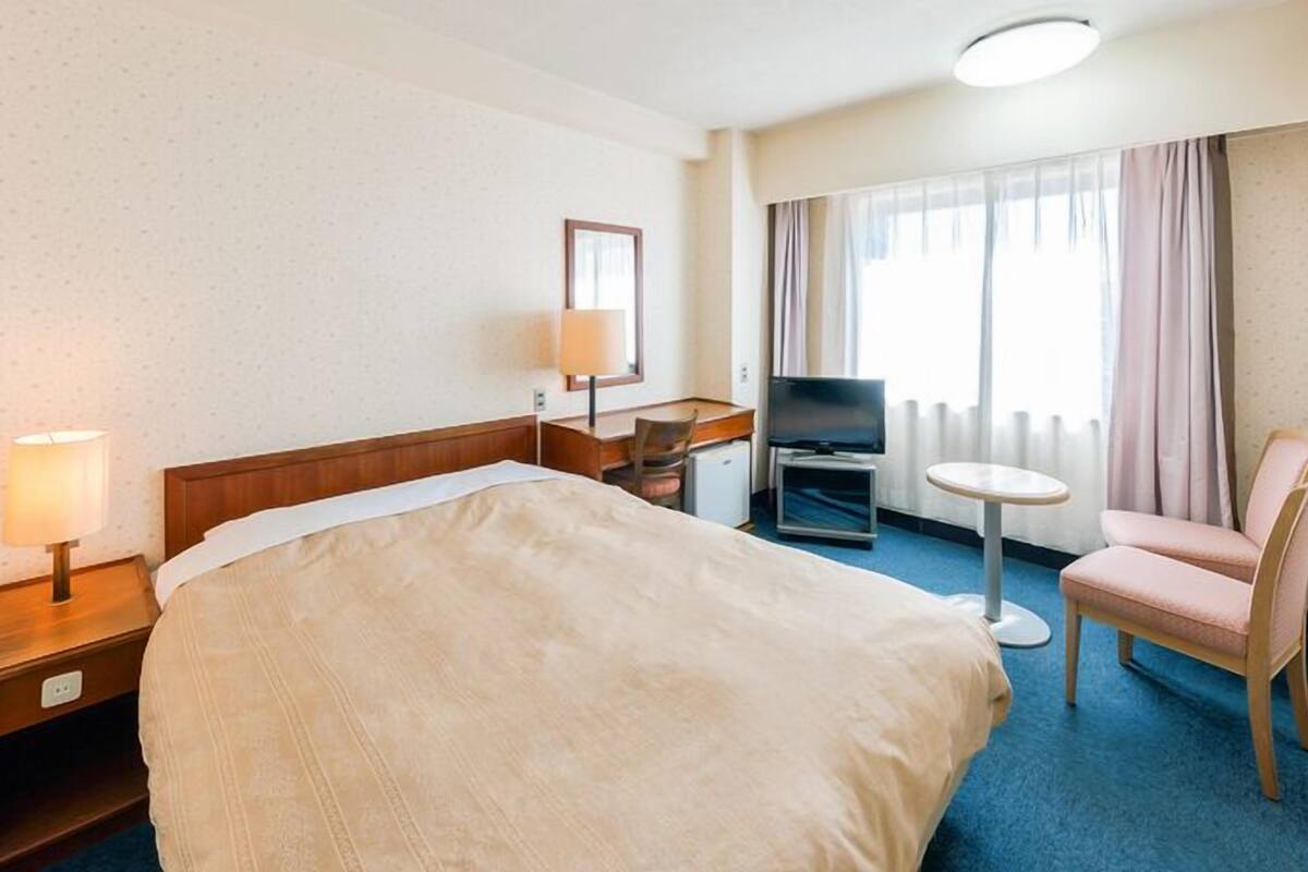 Ein Hotelzimmer mit Bett, Schreibtisch, Stuhl, Fernseher und einem Fenster mit Vorhängen.