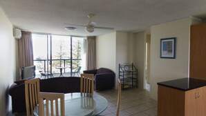A room with a round glass table, two chairs, a sofa, a TV, a ceiling fan, and a window with curtains. - Durham Court Holiday Apartments (Surfers Paradise)