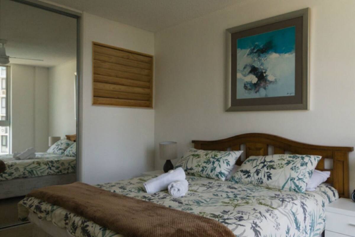 A hotel room with a bed, a mirror, a window with curtains, a painting on the wall, and a wooden headboard.