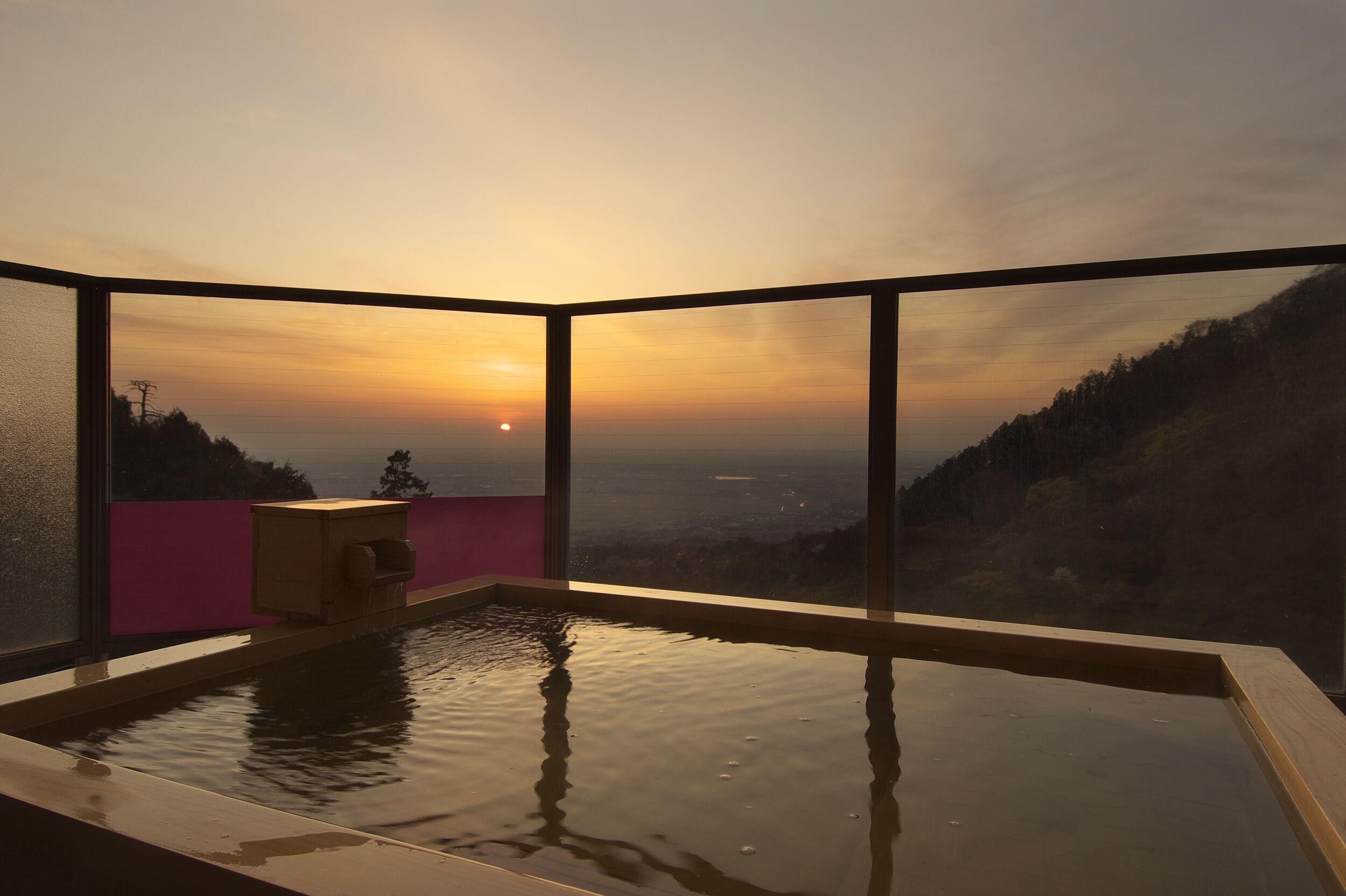Una habitación con un ventanal que da a un atardecer, un pequeño banco de madera y una fuente.