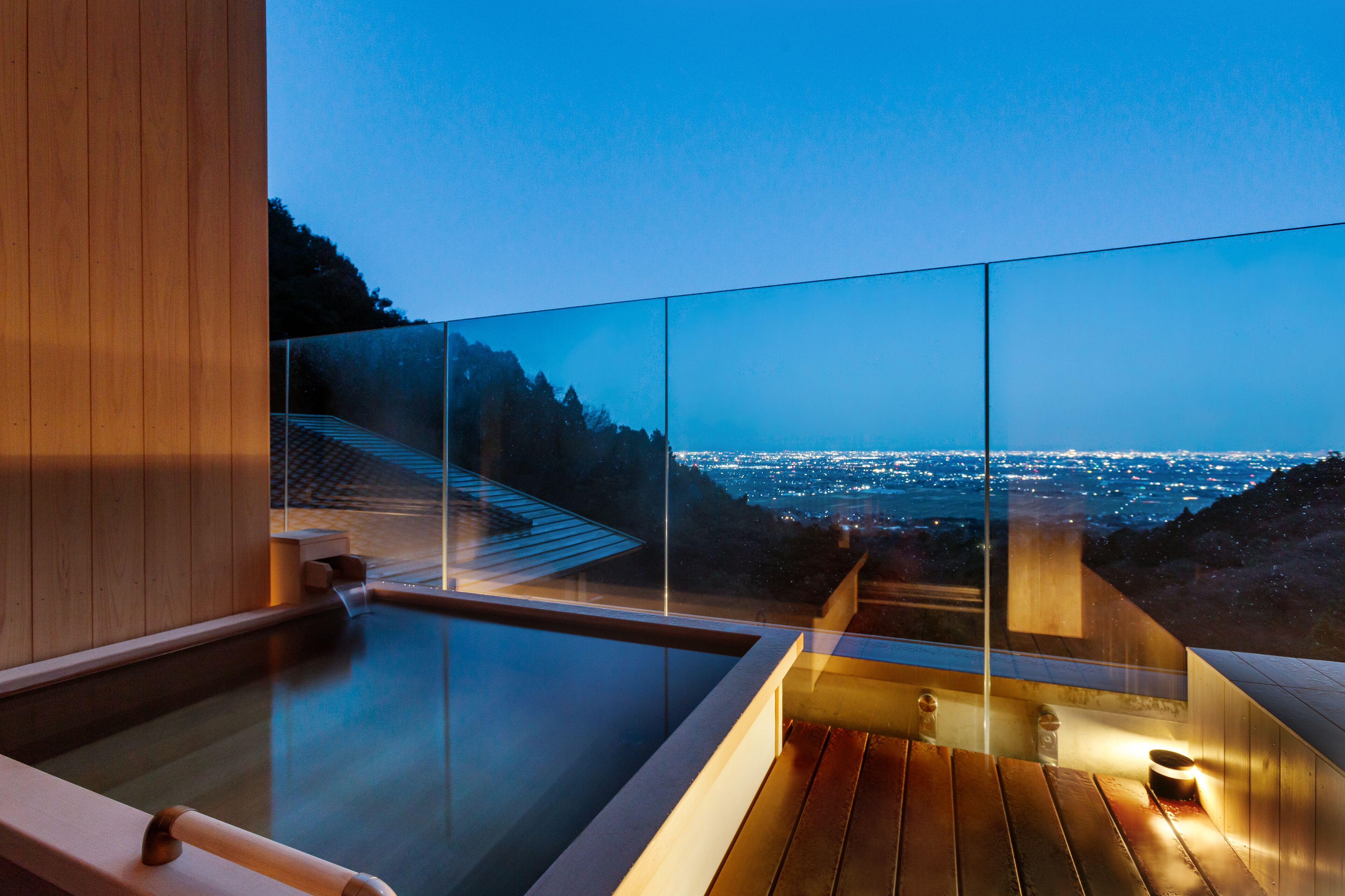 Una piscina moderna en la azotea con barandillas de cristal, terraza de madera y vistas a la ciudad al anochecer.
