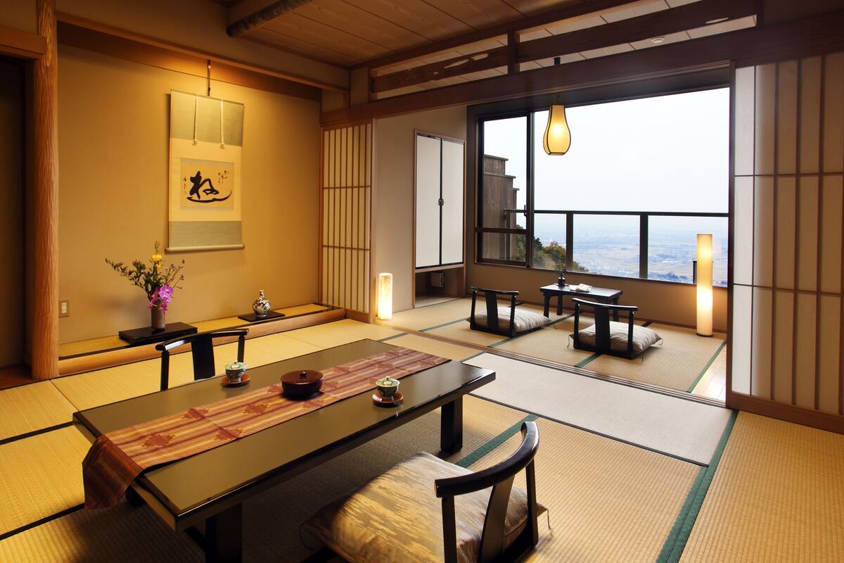 Une pièce de style japonais traditionnel, avec un sol recouvert de tatamis, une table basse avec des coussins, un luminaire suspendu et une vue sur le paysage urbain à travers de grandes fenêtres.