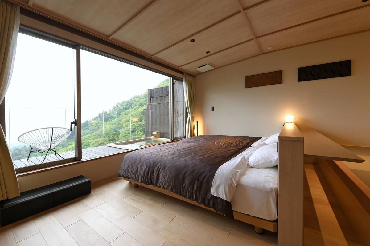 Une chambre avec une grande fenêtre, un lit, une table de chevet et une chaise.