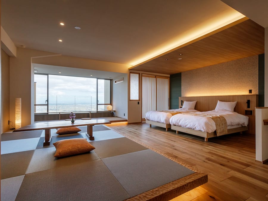 A modern hotel room with a large bed, a seating area with tatami mats, a view of the city, and a wooden ceiling.
