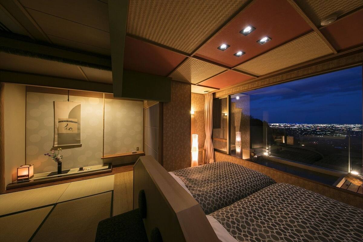 Une chambre d’hôtel avec une grande fenêtre, un lit, une coiffeuse avec un miroir et une petite table sur laquelle est posé un vase.