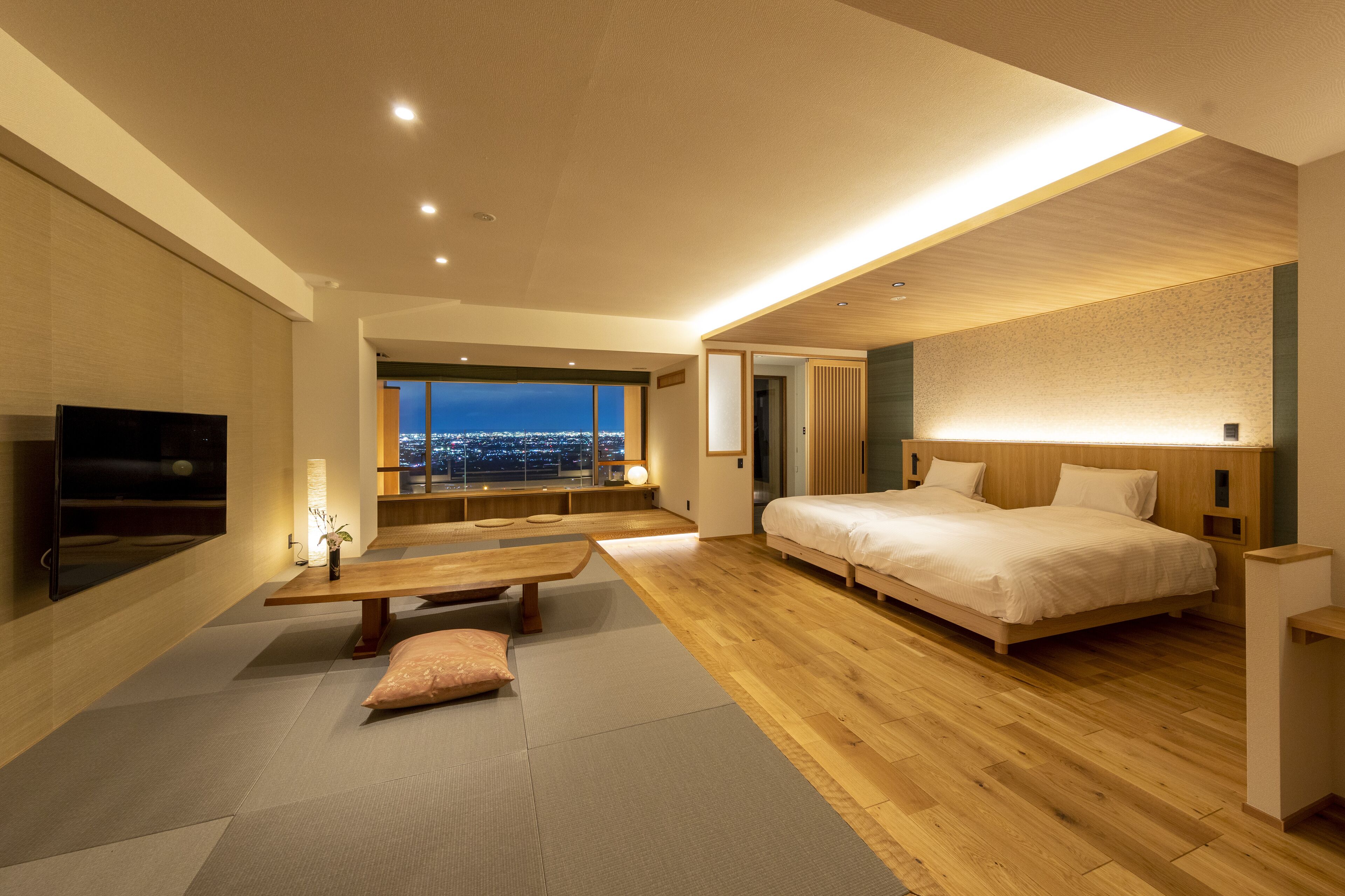 A modern hotel room with a large bed, a TV, a wooden bench, and a view of the city at night.