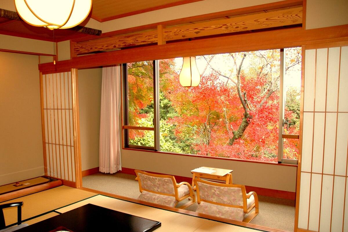 Une pièce de style japonais traditionnel, avec un sol recouvert de tatamis, une table basse, deux chaises, une fenêtre donnant sur le feuillage automnal et un luminaire suspendu.
