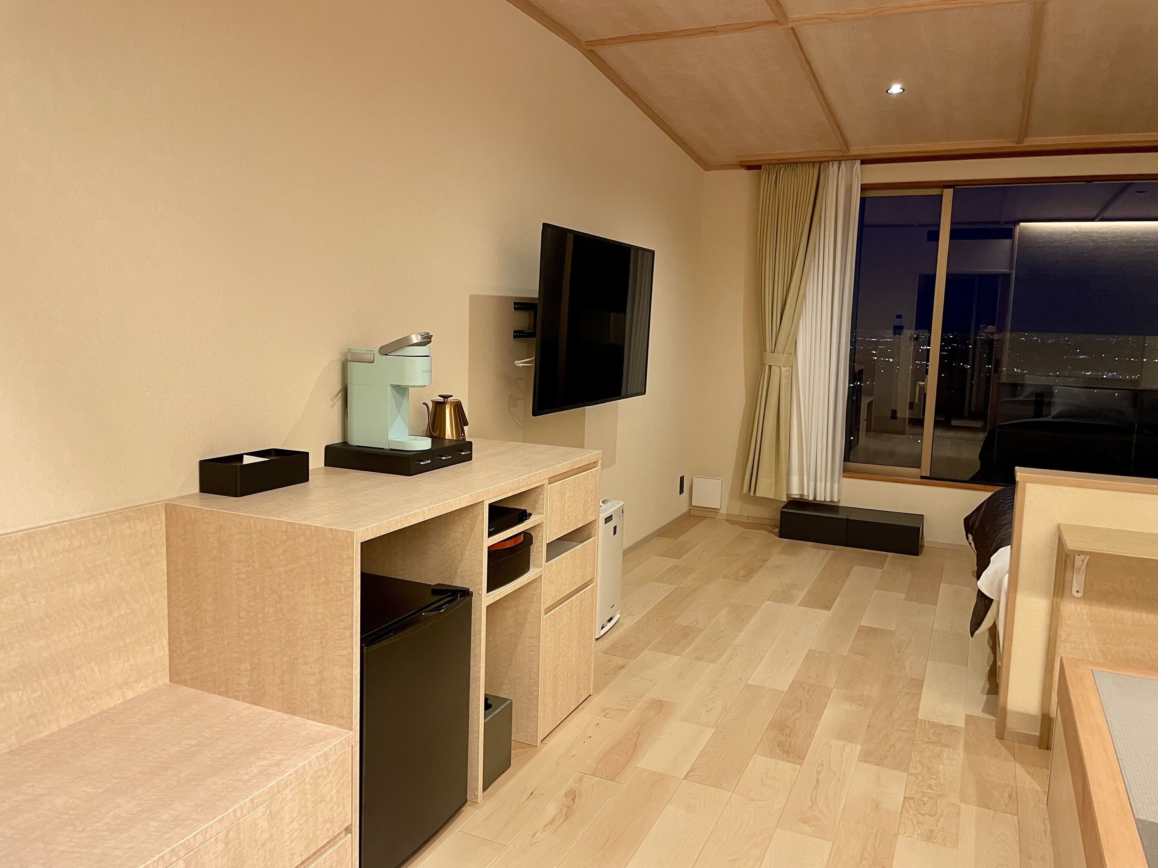 A modern hotel room with a wooden desk, a flat-screen TV, a coffee maker, and a city view through large windows.