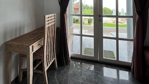 Una habitación con un escritorio de madera, una silla y un amplio ventanal corredera que da a un área exterior con sombrillas.