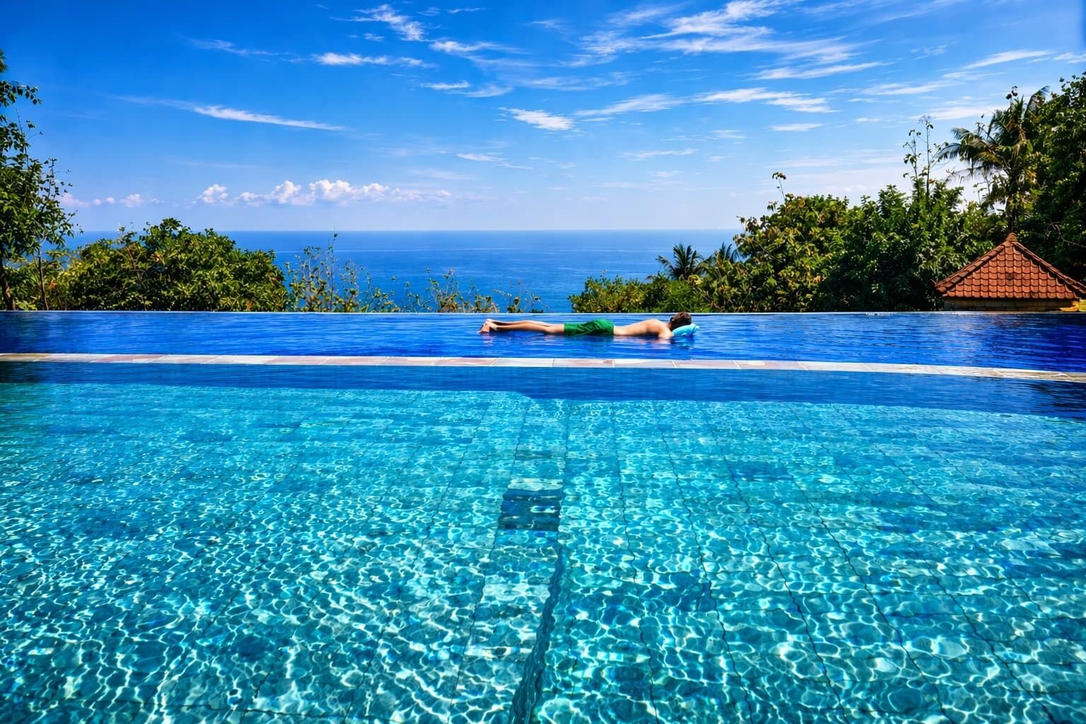 A clear blue infinity pool with a person lying on the edge, surrounded by lush greenery and a view of the ocean.