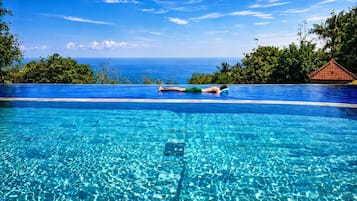 Una piscina infinita de color azul intenso con una persona recostada en el borde, rodeada de exuberante vegetación y vistas al océano.