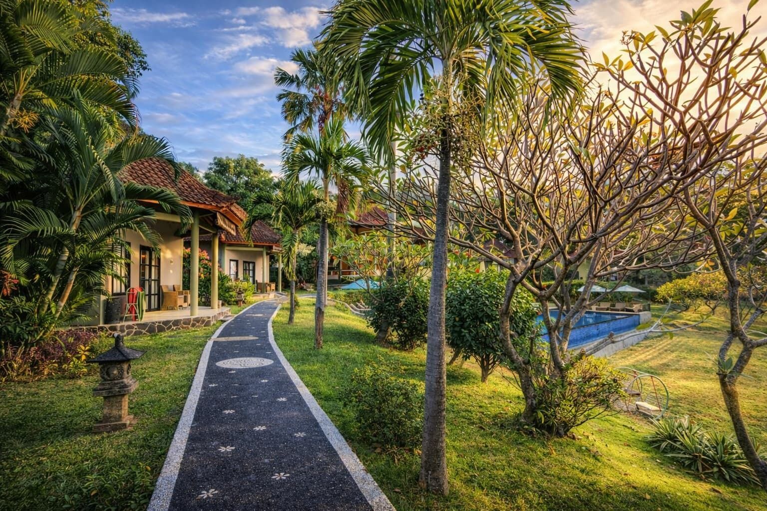 A pathway leading to a building with a tiled roof, surrounded by lush greenery and a swimming pool.