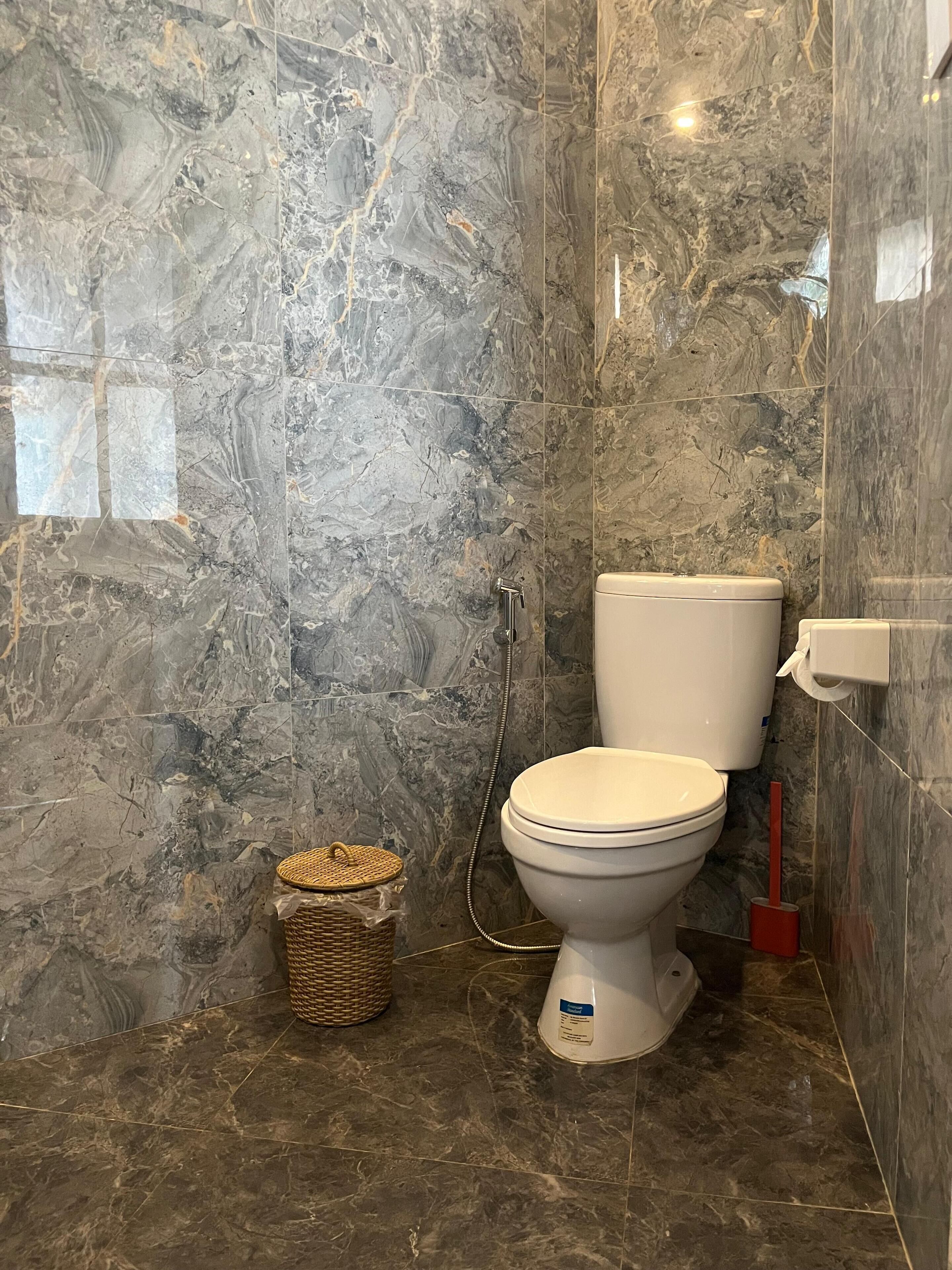 Ein Badezimmer mit Marmorboden und -wänden, einer weißen Toilette, einem Korbwannenstuhl und einem roten Pömpel.