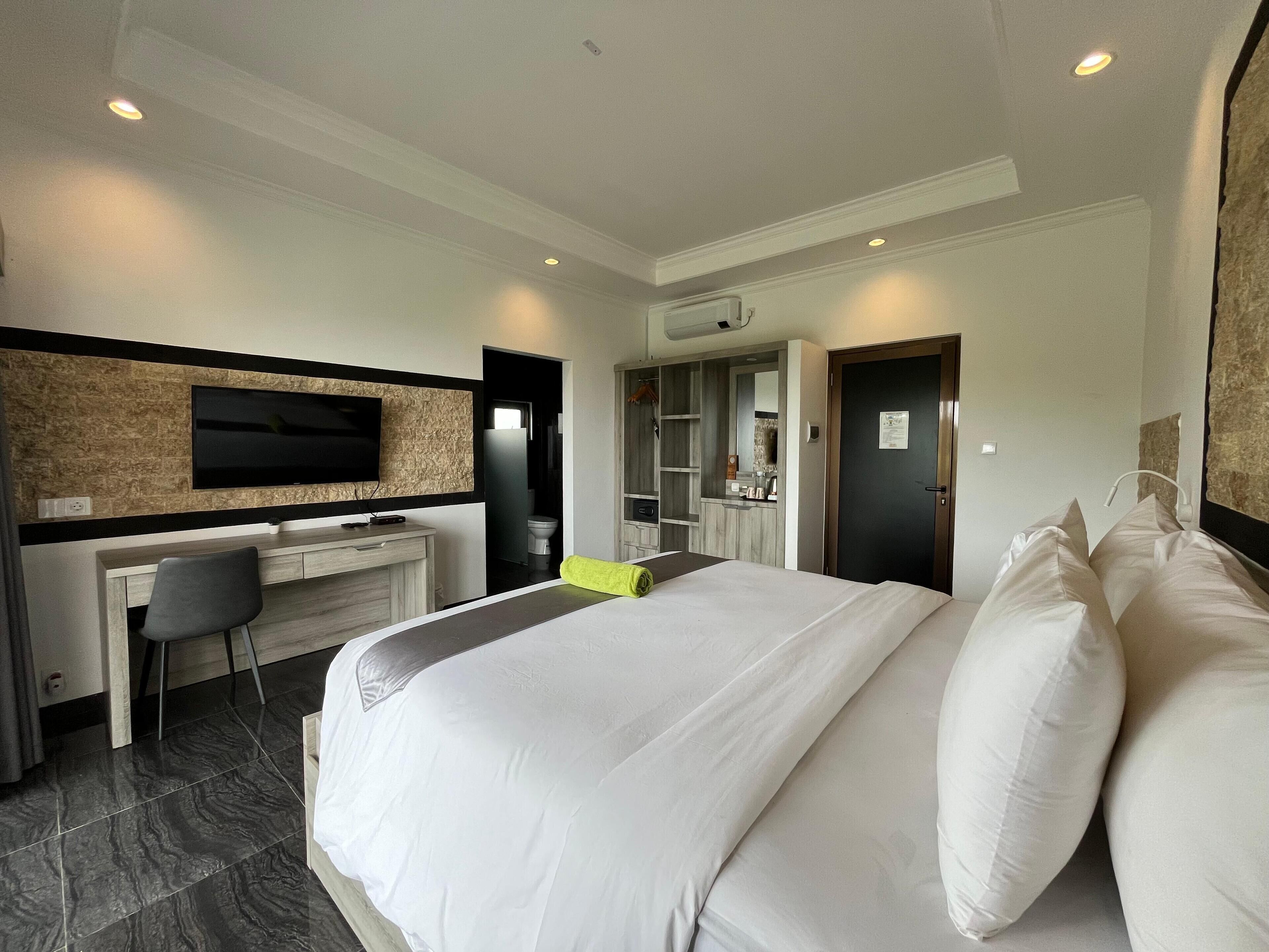 Une chambre d’hôtel moderne, dotée d’un grand lit, d’un bureau avec une chaise, d’un téléviseur à écran plat fixé au mur et d’une salle de bain visible à travers une porte ouverte.