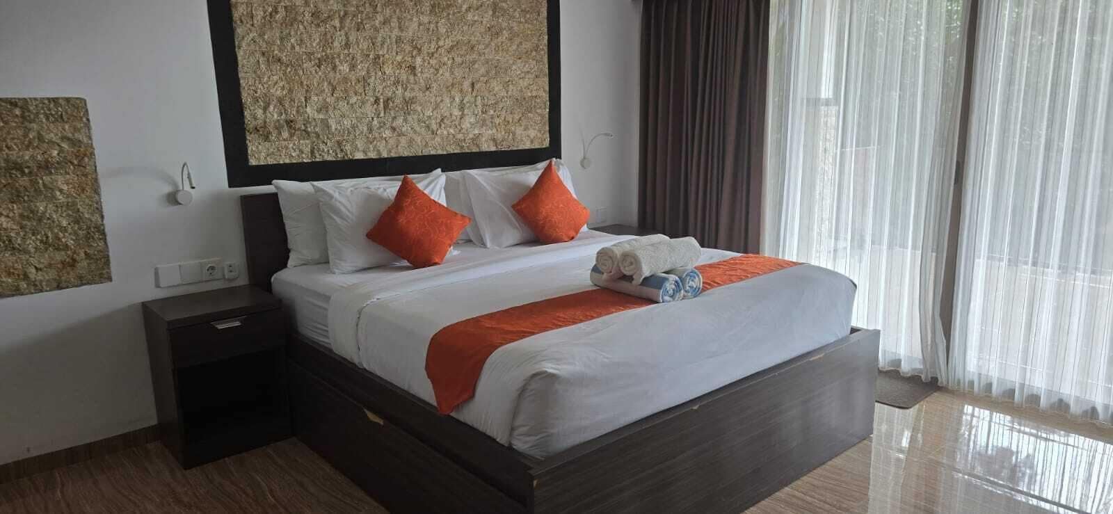 A hotel room with a large bed, a nightstand, a wall-mounted artwork, and a view of the outdoors through sheer curtains.