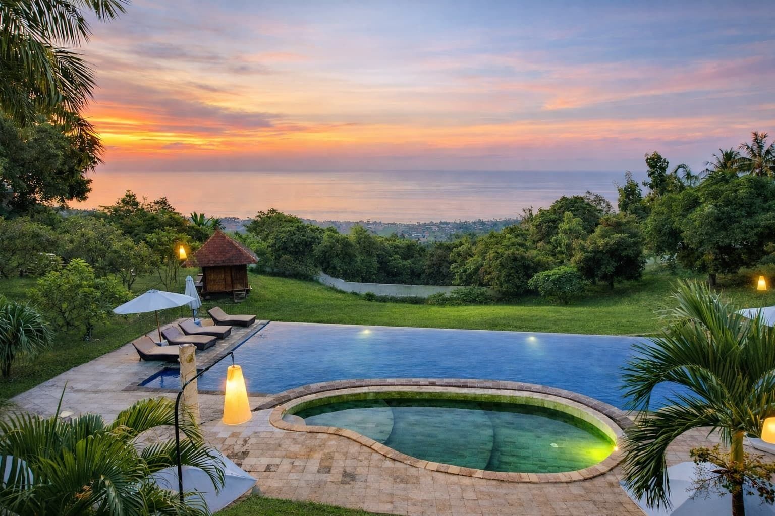 A swimming pool with a curved edge, surrounded by lounge chairs and umbrellas, set against a backdrop of a sunset and lush greenery.