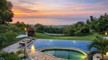 A swimming pool with a curved edge, surrounded by lounge chairs and umbrellas, set against a backdrop of a sunset and lush greenery.