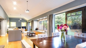 A spacious living area with a dining table, comfortable seating, a kitchen in the background, and large windows offering a view of the outdoors. - Parehua Resort (Martinborough)
