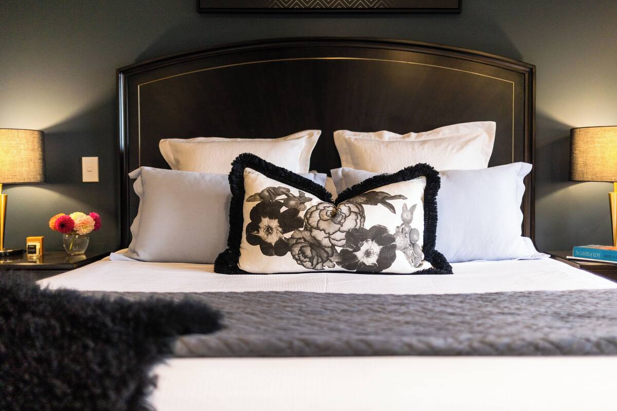 A neatly made bed with white linens and decorative pillows, including one with a floral design. There are two bedside lamps on either side of the bed, and a small vase with flowers on the nightstand.