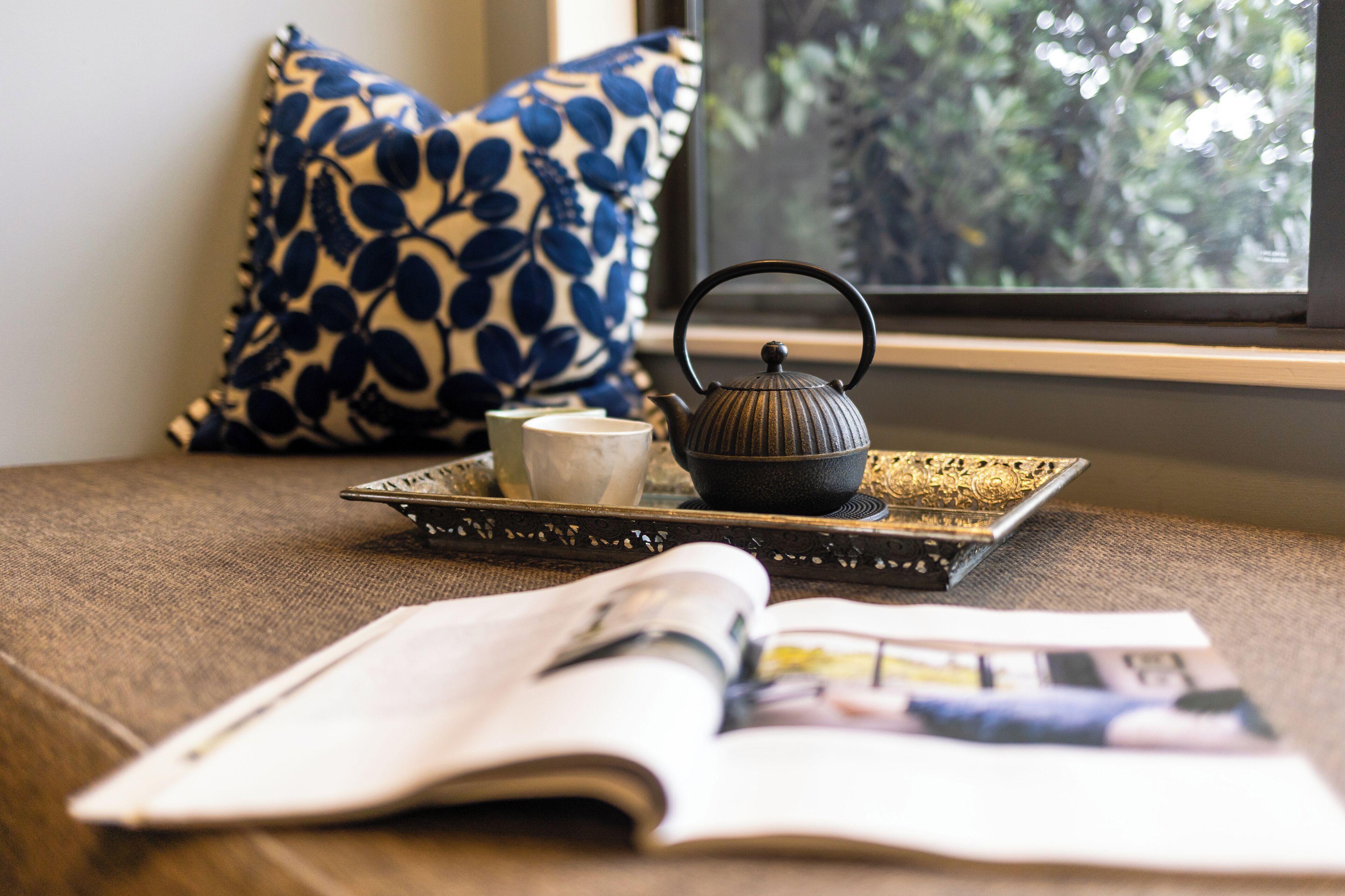 Une pièce avec une fenêtre, un coussin à motifs, un plateau avec une théière et une tasse, et un magazine ouvert.