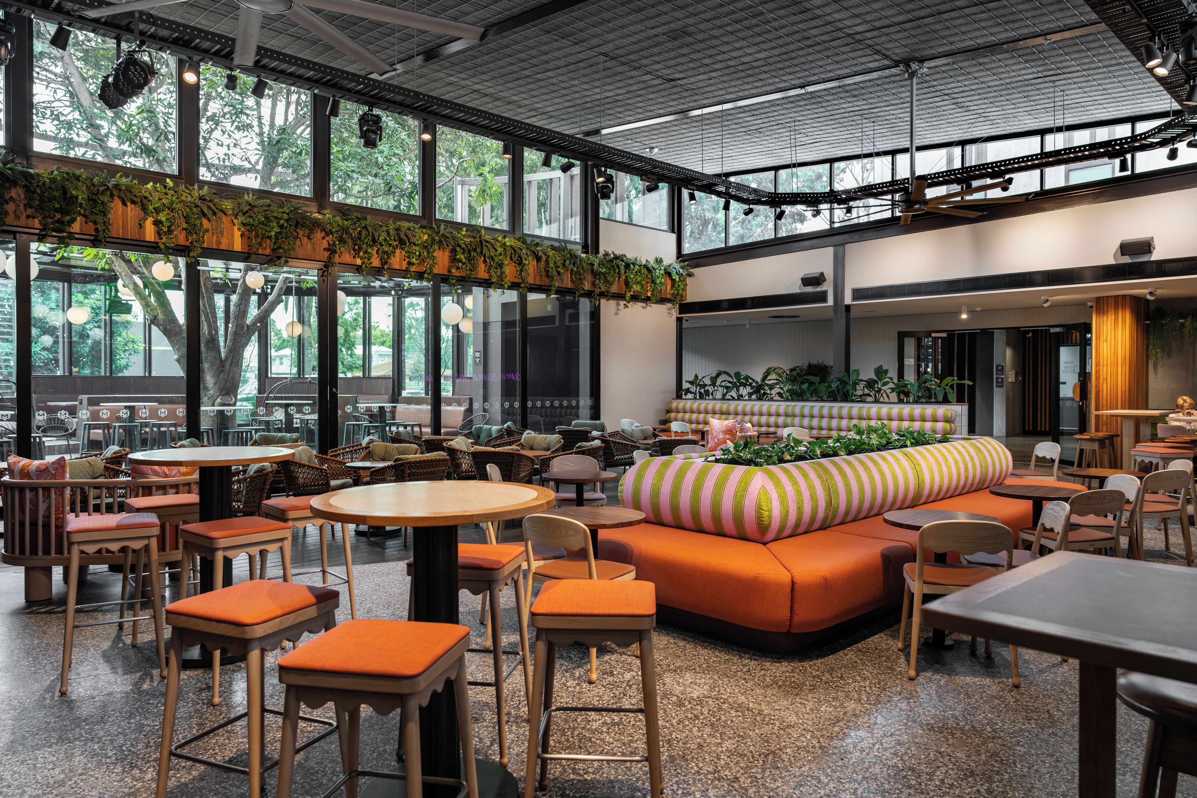 A spacious indoor area with multiple seating arrangements including tables and sofas, large windows providing natural light, and a ceiling with exposed beams.
