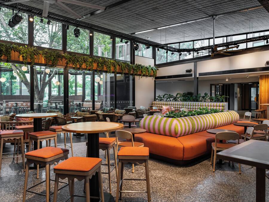 A spacious indoor area with multiple seating arrangements including tables and sofas, large windows providing natural light, and a ceiling with exposed beams.