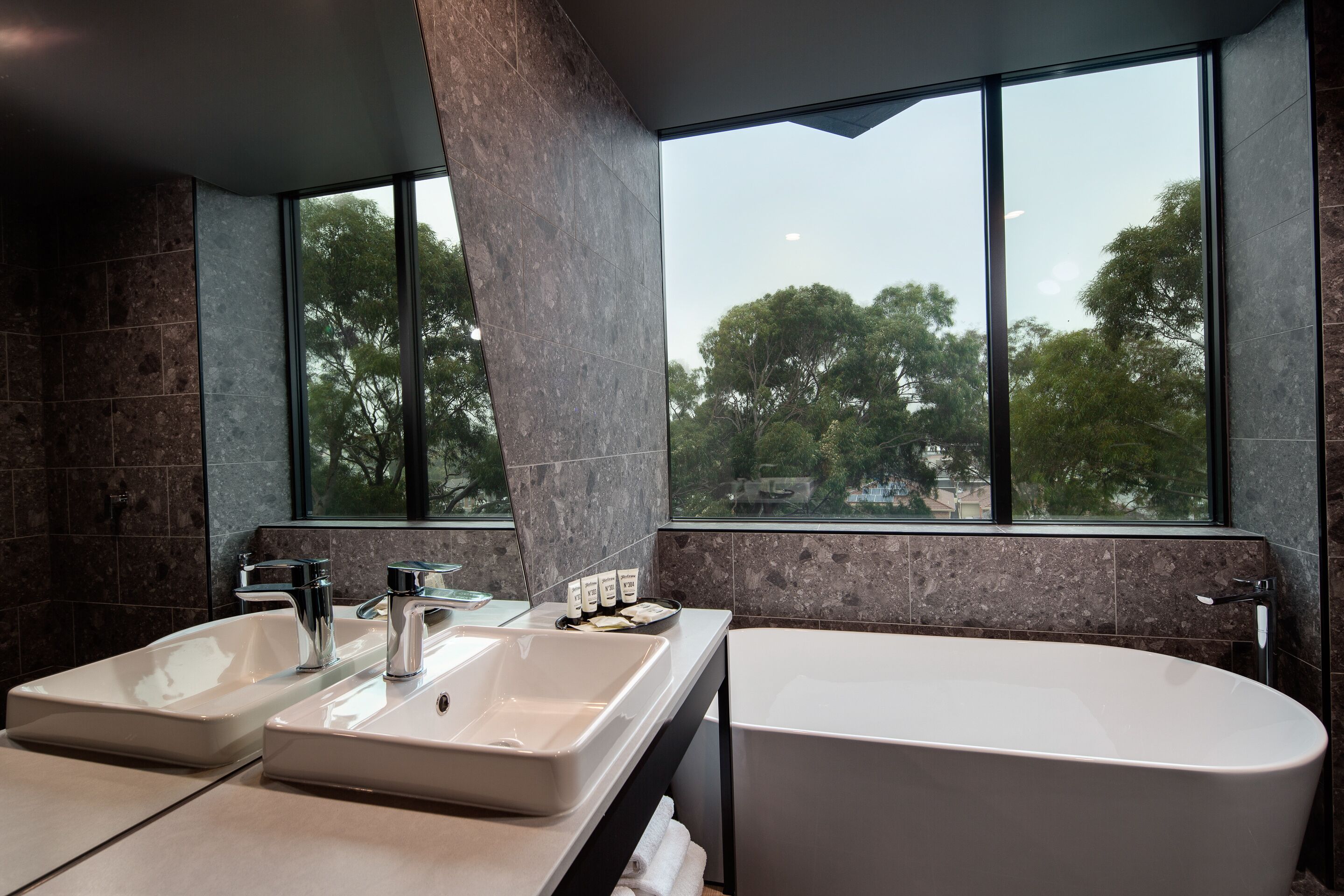 Un baño moderno con dos lavamanos, una bañera y un amplio ventanal con vistas a árboles.