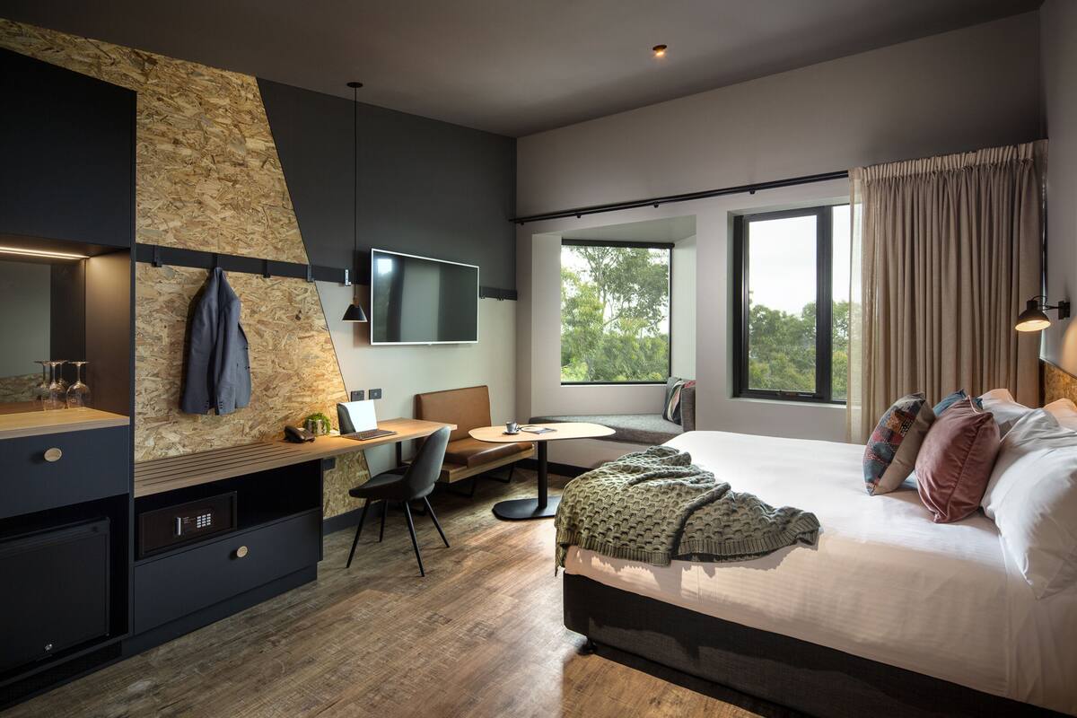 A modern hotel room with a bed, a desk, a chair, a small table, and a large window.