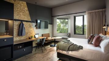 Una habitación de hotel moderna con cama, escritorio, silla, mesita y un gran ventanal.