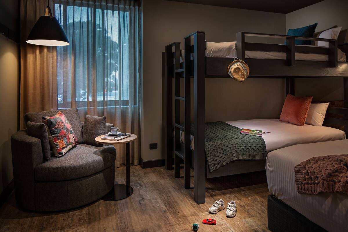 A compact hotel room with a bunk bed, a sofa, a small table, and a window with curtains.