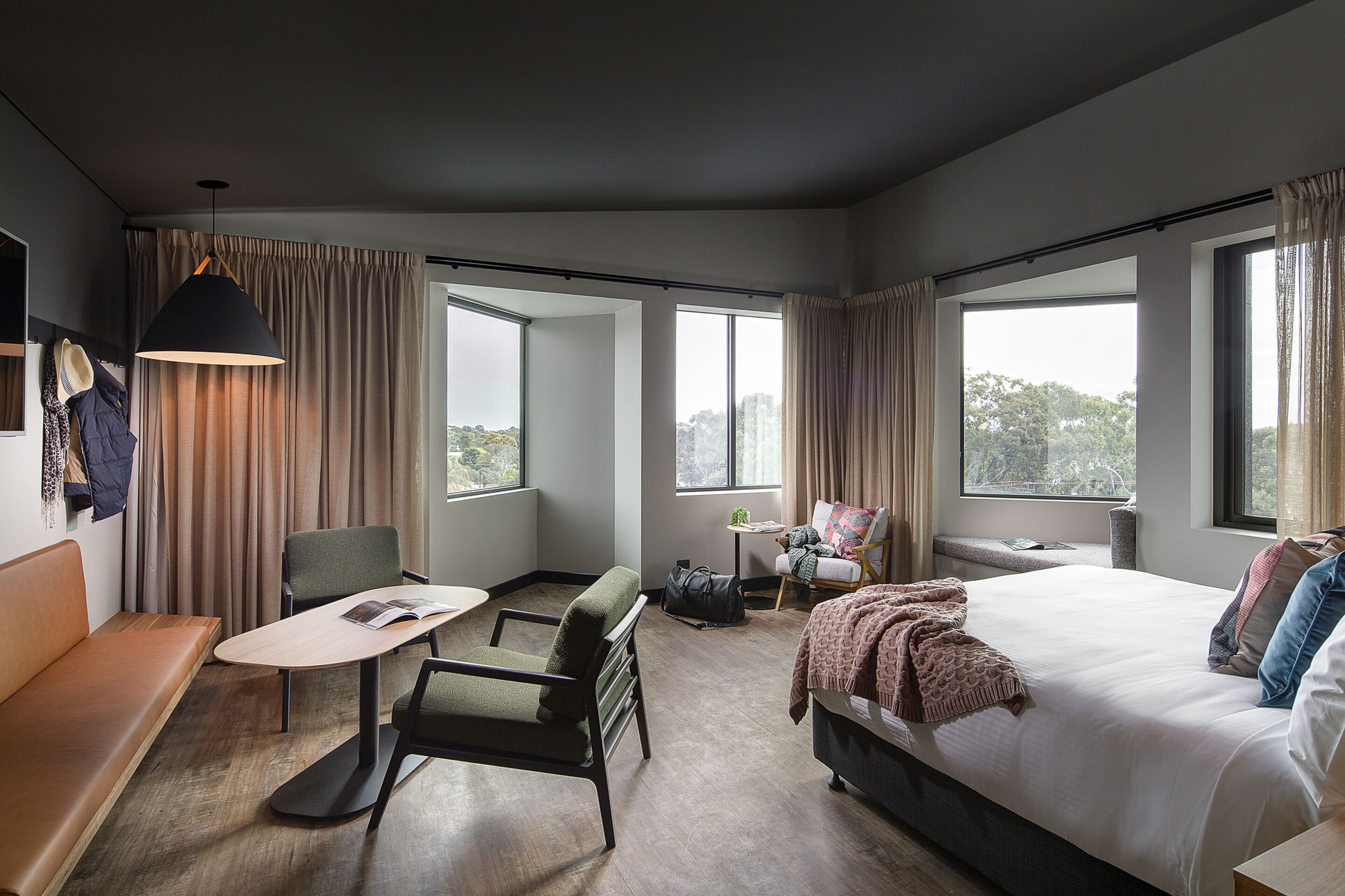 A modern hotel room with a large bed, a sofa, a dining table, and a chair. There are large windows with curtains, a hanging light fixture, and a view of the outdoors.