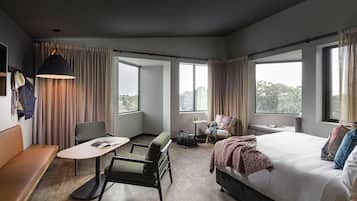 A modern hotel room with a large bed, a sofa, a dining table, and a chair. There are large windows with curtains, a hanging light fixture, and a view of the outdoors.