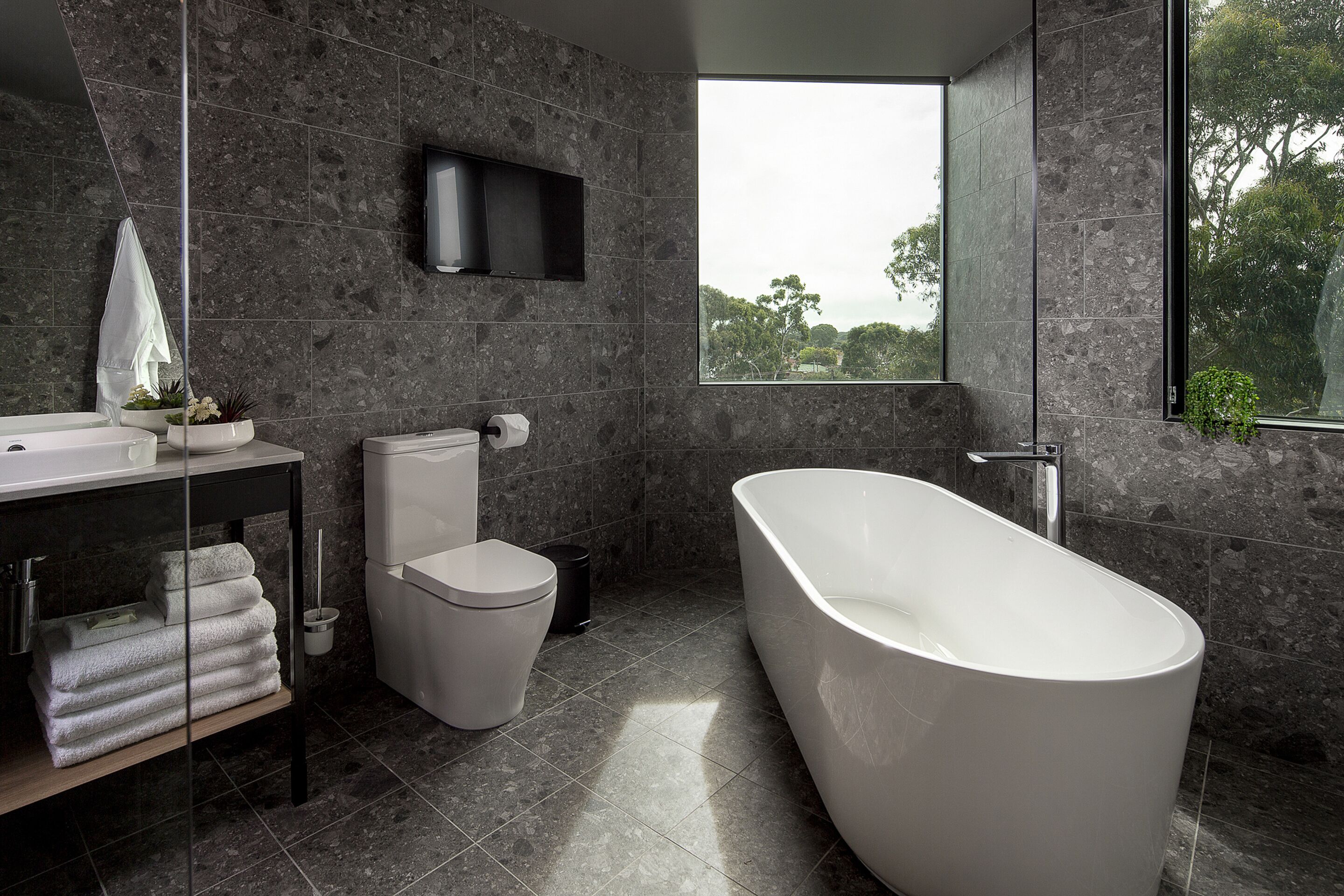 A modern bathroom with a freestanding bathtub, a toilet, a sink, and a large window offering a view of trees.