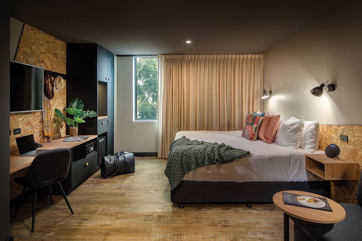 A modern hotel room with a wooden desk, a bed with multiple pillows, a bedside table with a lamp, and a window with curtains.