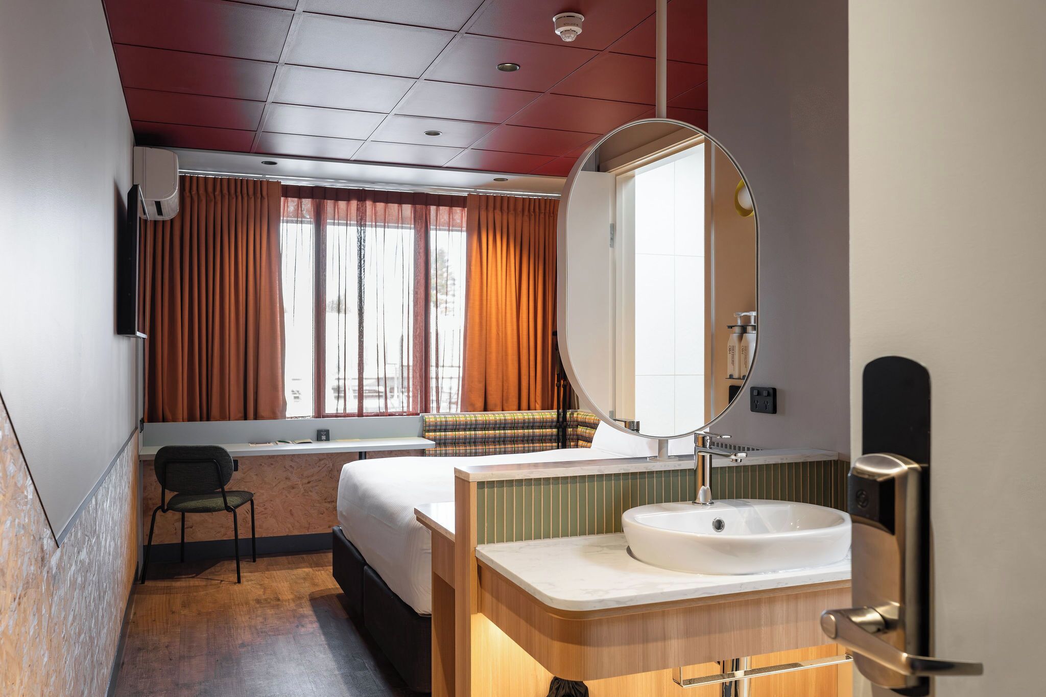 A hotel room with a bed, a desk, a chair, a large mirror, a sink, and a window with curtains.