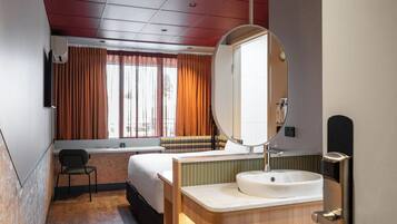 A hotel room with a bed, a desk, a chair, a large mirror, a sink, and a window with curtains.