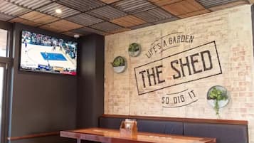 A restaurant interior with a brick wall, a mounted TV, and a slogan 'LIFE'S A GARDEN THE SHED SO DIG IT'.