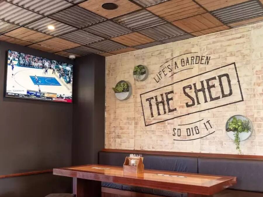A restaurant interior with a brick wall, a mounted TV, and a slogan 'LIFE'S A GARDEN THE SHED SO DIG IT'.