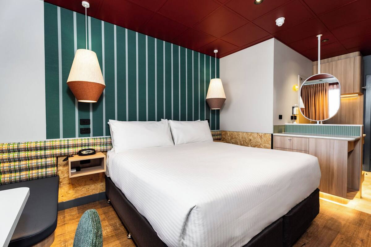 A modern hotel room with a large bed, a small desk, a chair, a nightstand, a mirror, and a wall with vertical stripes.