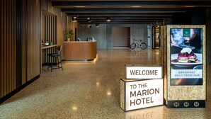 Zona de recepción de un hotel con un cartel de bienvenida, mostrador de recepción, una bicicleta y una pantalla digital que anuncia el desayuno.