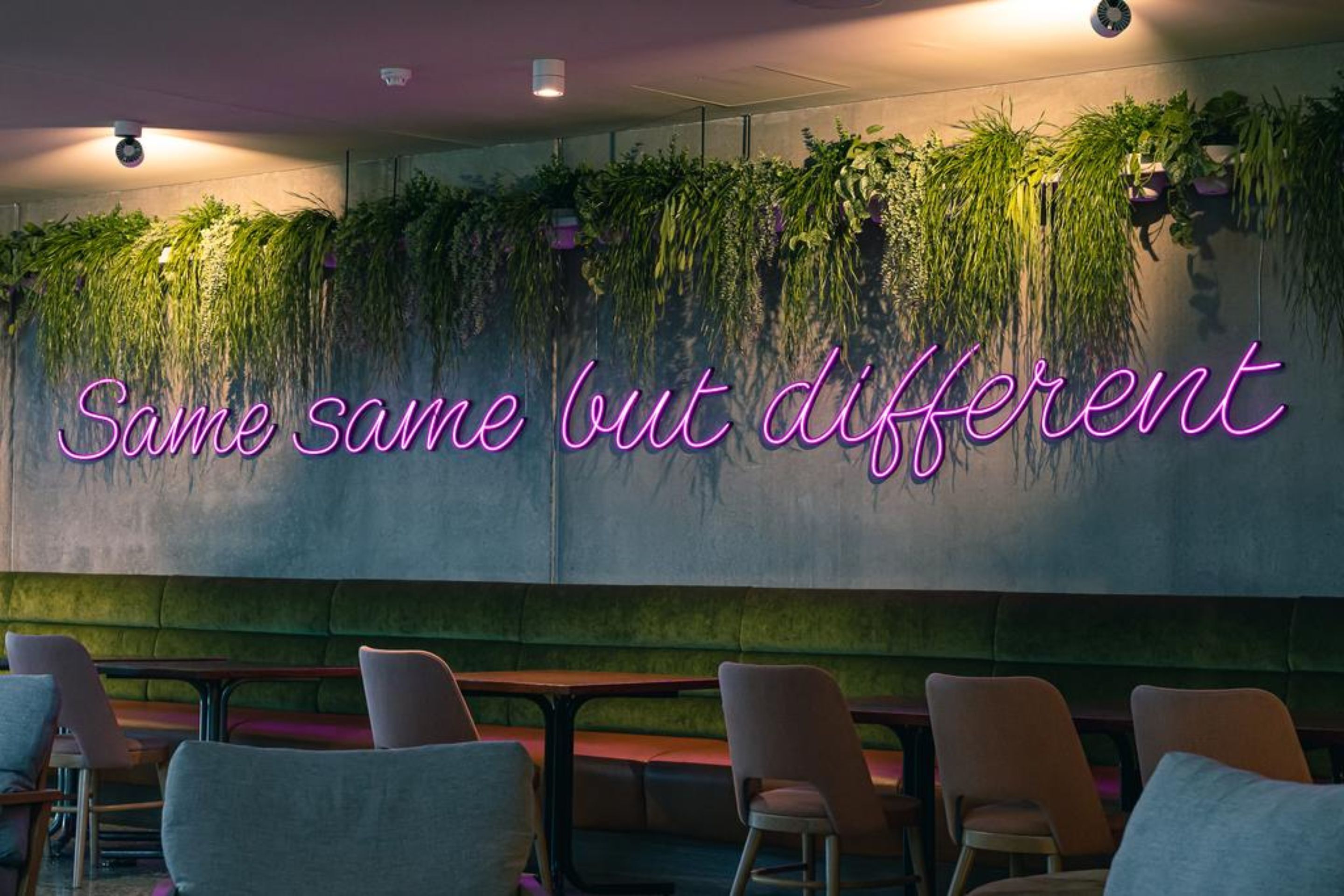 A restaurant interior with a neon sign reading "Same same but different" and a wall of hanging plants.